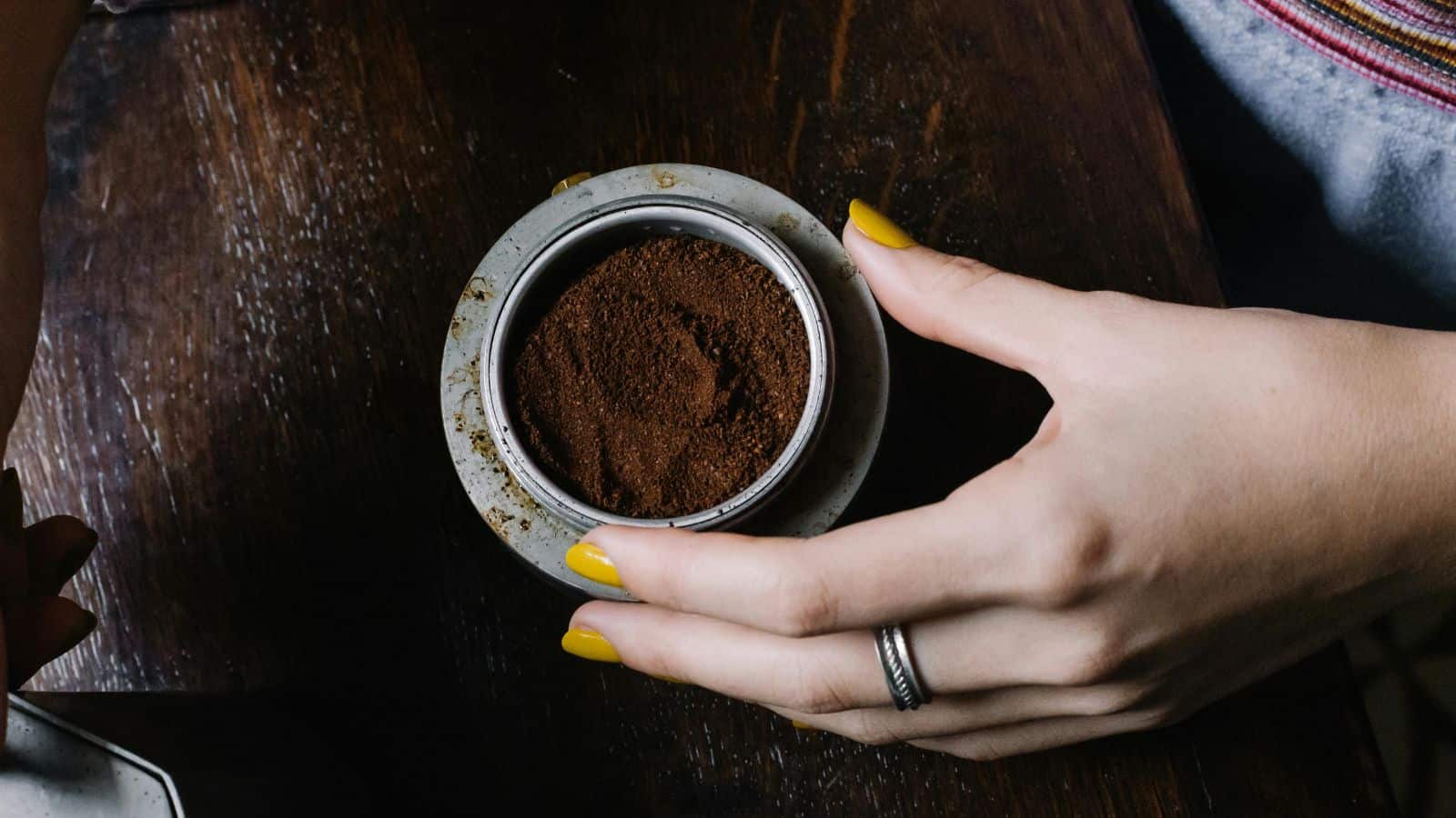 A person with yellow-painted nails holds a portafilter filled with ground coffee over a dark wooden surface.