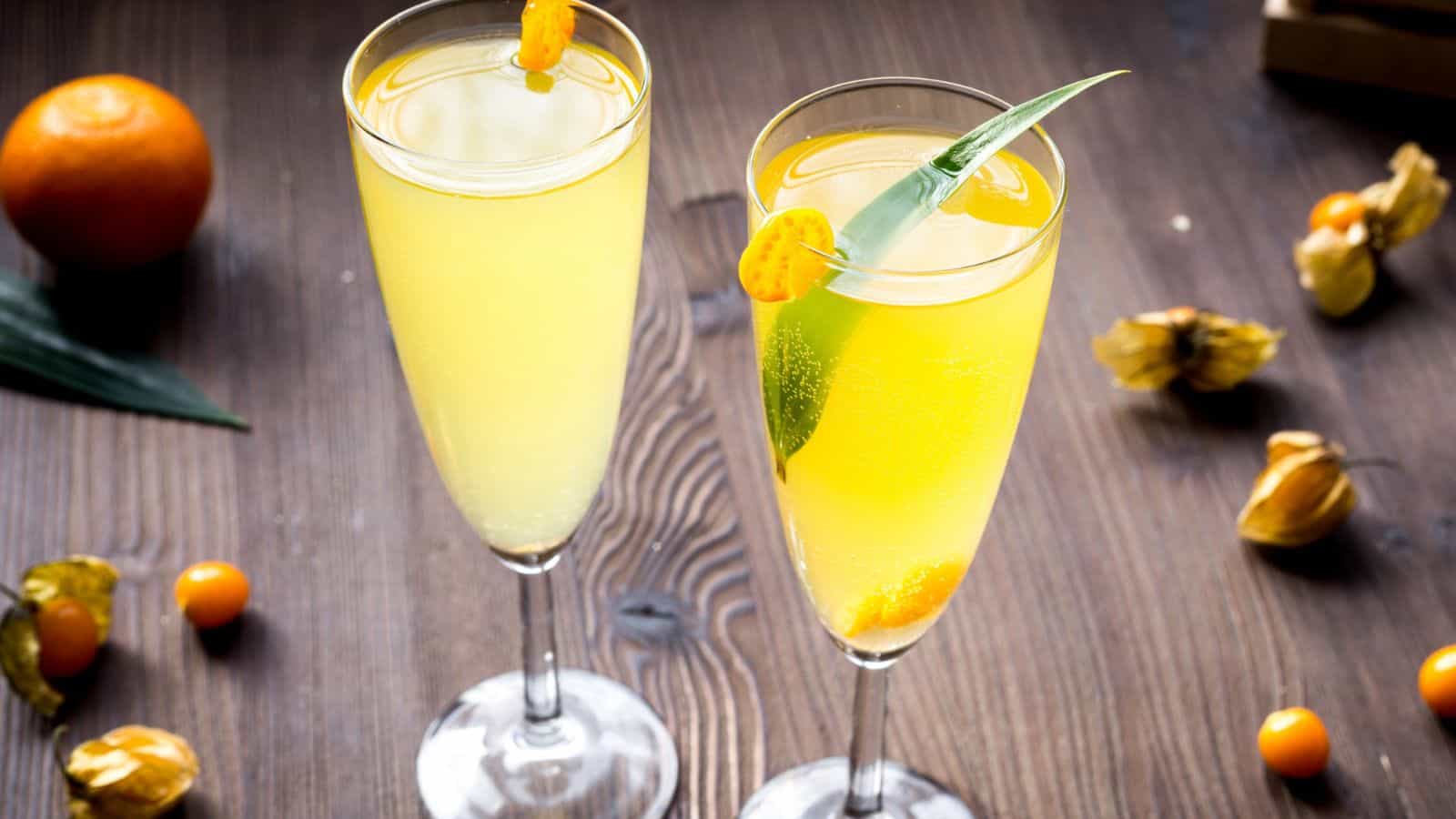 Two tall glasses filled with a yellow cocktail are garnished with a small orange fruit and a green leaf. The drinks are placed on a wooden table surrounded by scattered fruits and leaves. A whole orange can be seen in the background.