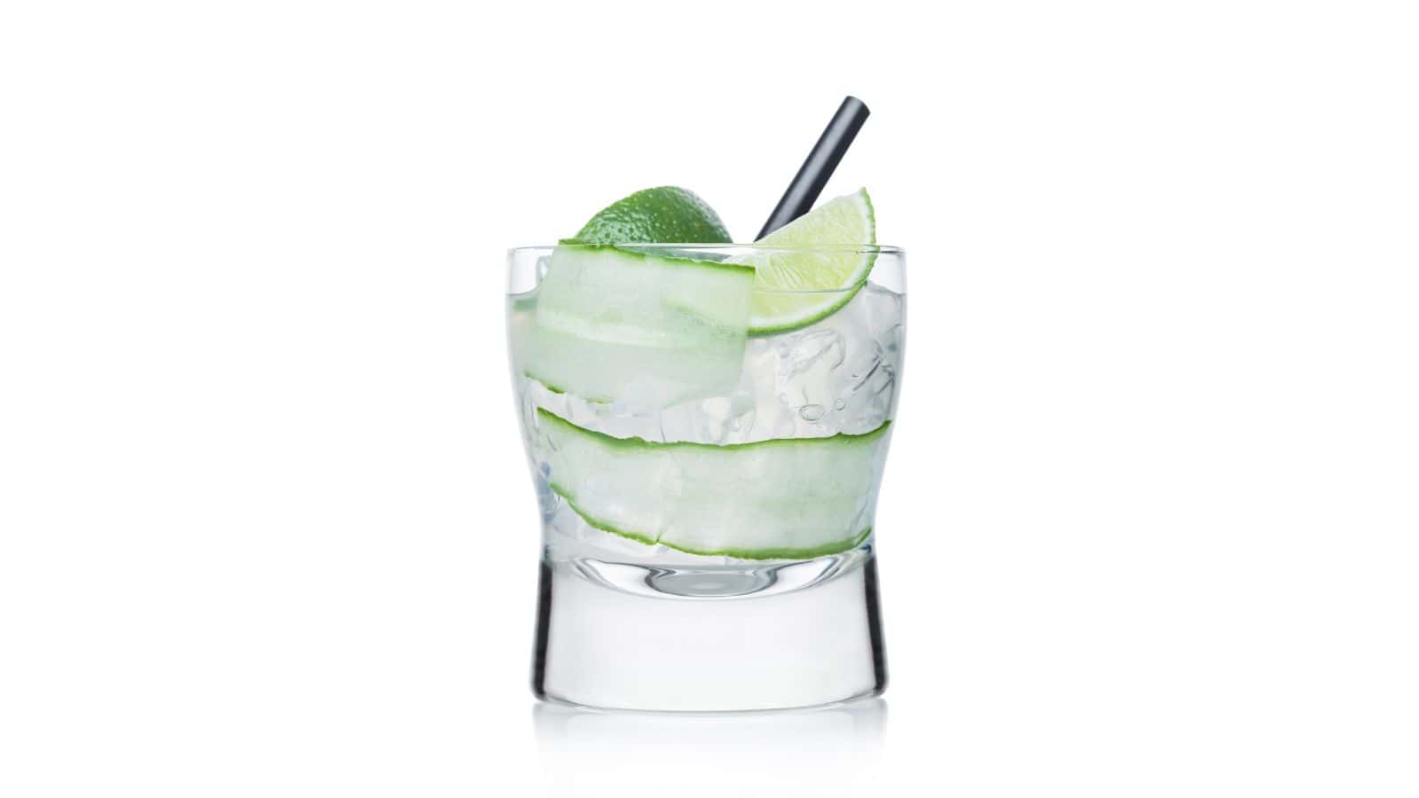 A clear glass filled with a transparent liquid, large ice cubes, cucumber slices, and a lime wedge on the rim. A black straw is placed inside. The glass is on a white background.
