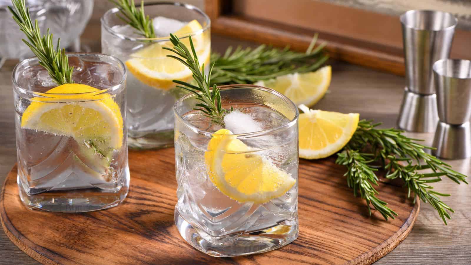 Three glasses of lemon and rosemary gin and tonic with ice are on a wooden board. Slices of lemon and sprigs of rosemary are also on the board. Metal measuring jiggers are visible in the background.