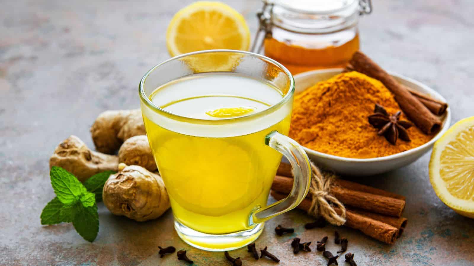 A glass cup of yellow ginger tea is surrounded by ingredients including fresh ginger, lemon slices, a bowl of turmeric powder, a jar of honey, cinnamon sticks, star anise, cloves, and mint leaves.