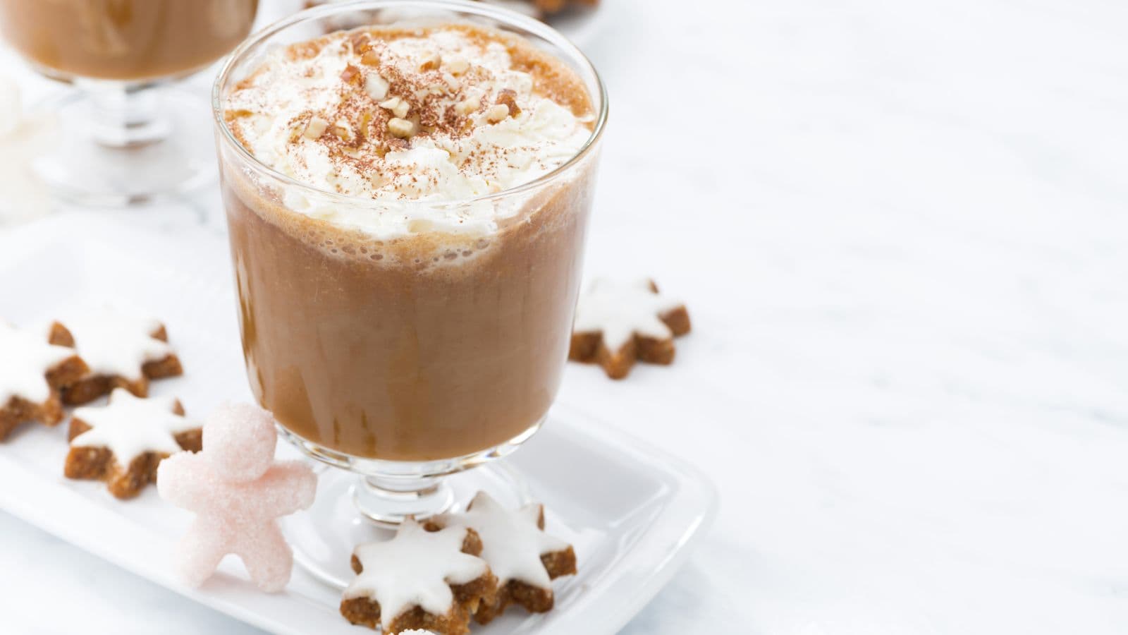 A glass of frothy hot chocolate topped with whipped cream and sprinkled with crushed nuts is on a white tray. Surrounding the glass are star-shaped cookies and a small sugar-coated figure-shaped treat.