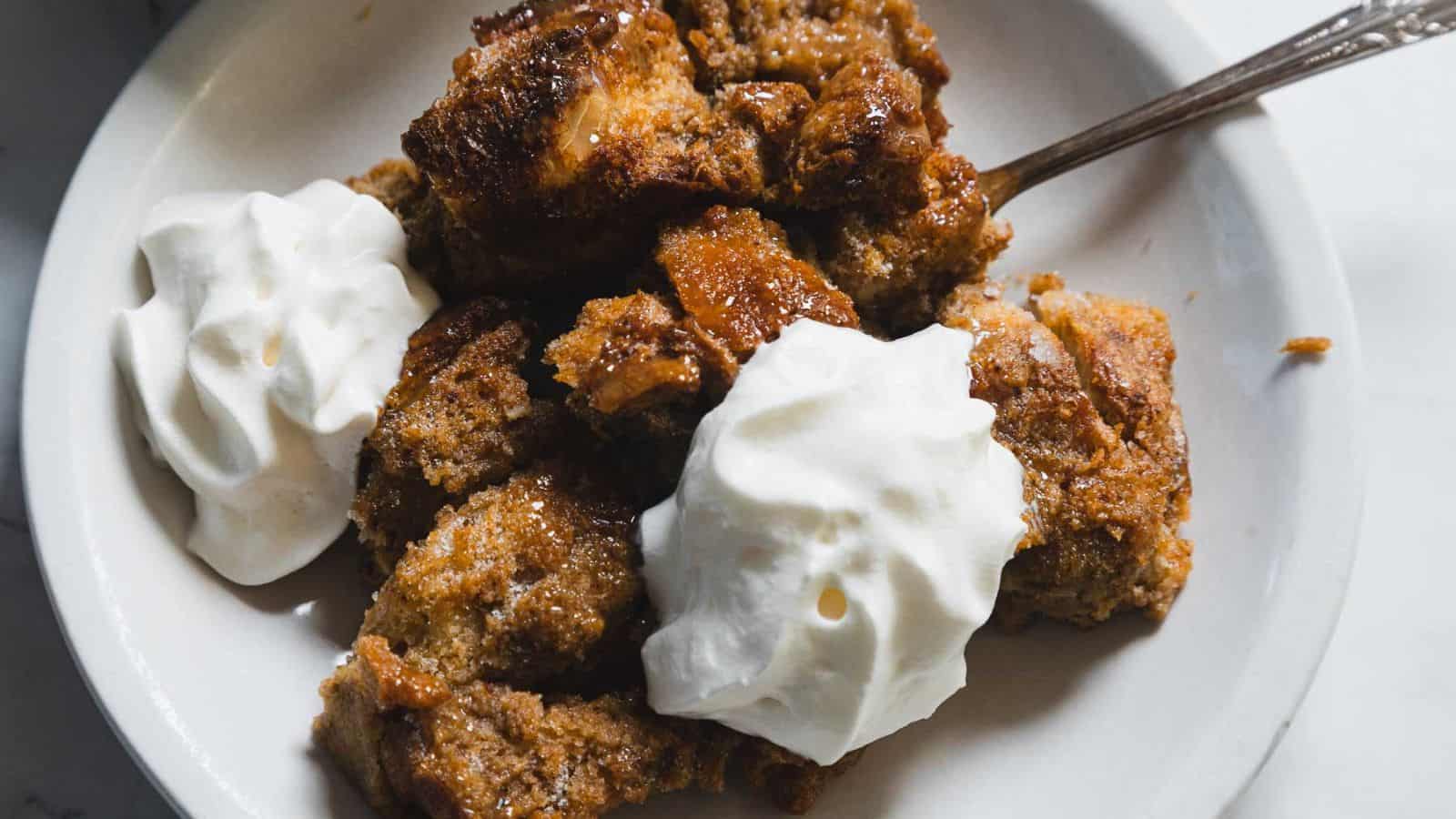 A plate of bread pudding topped with two dollops of whipped cream. A fork rests on the plate. In the background, a baking dish with more bread pudding is partially visible on a marble surface. Decorative items are placed around.