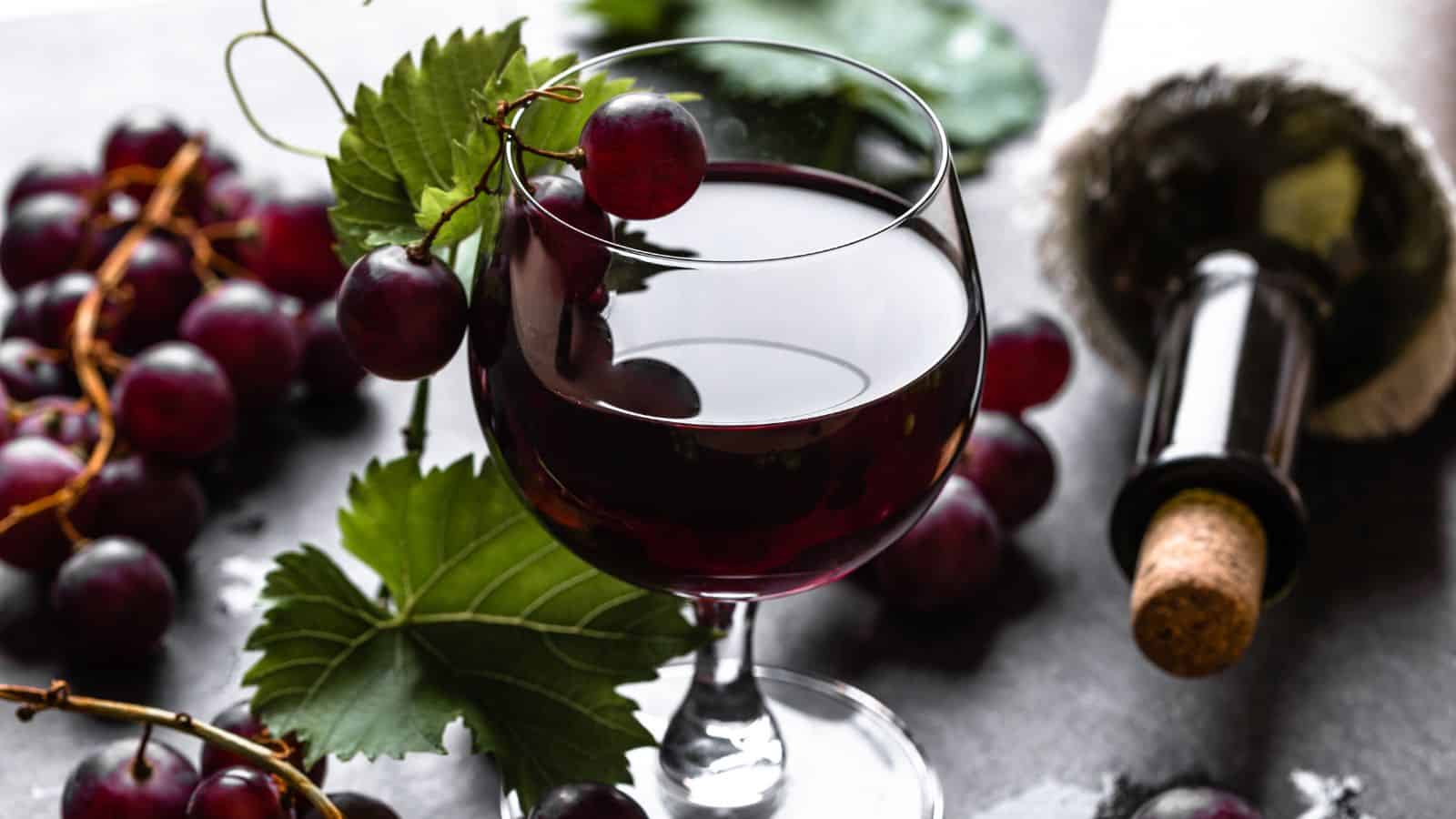 A glass of red wine is placed on a dark surface. Surrounding the glass are several bunches of red grapes and green leaves. A wine bottle with a cork rests nearby, tilted on its side, partially out of focus.