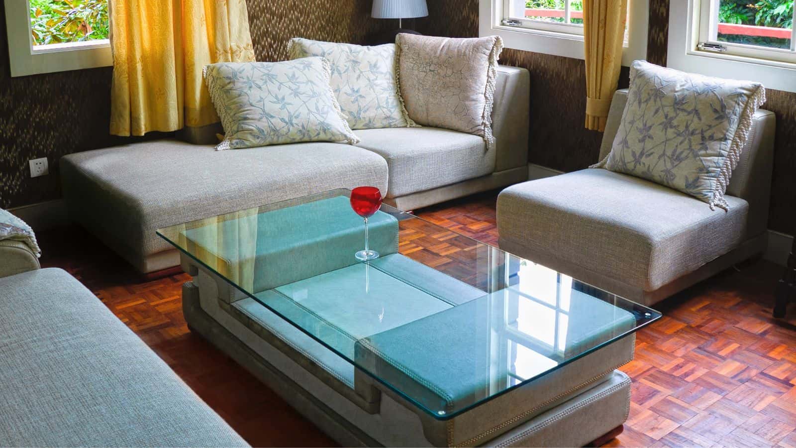 A living room features a glass coffee table with a single red wine glass on it, surrounded by light gray sectional sofas and plush pillows. The hardwood floor complements the room's warm tones, while windows let in natural light.