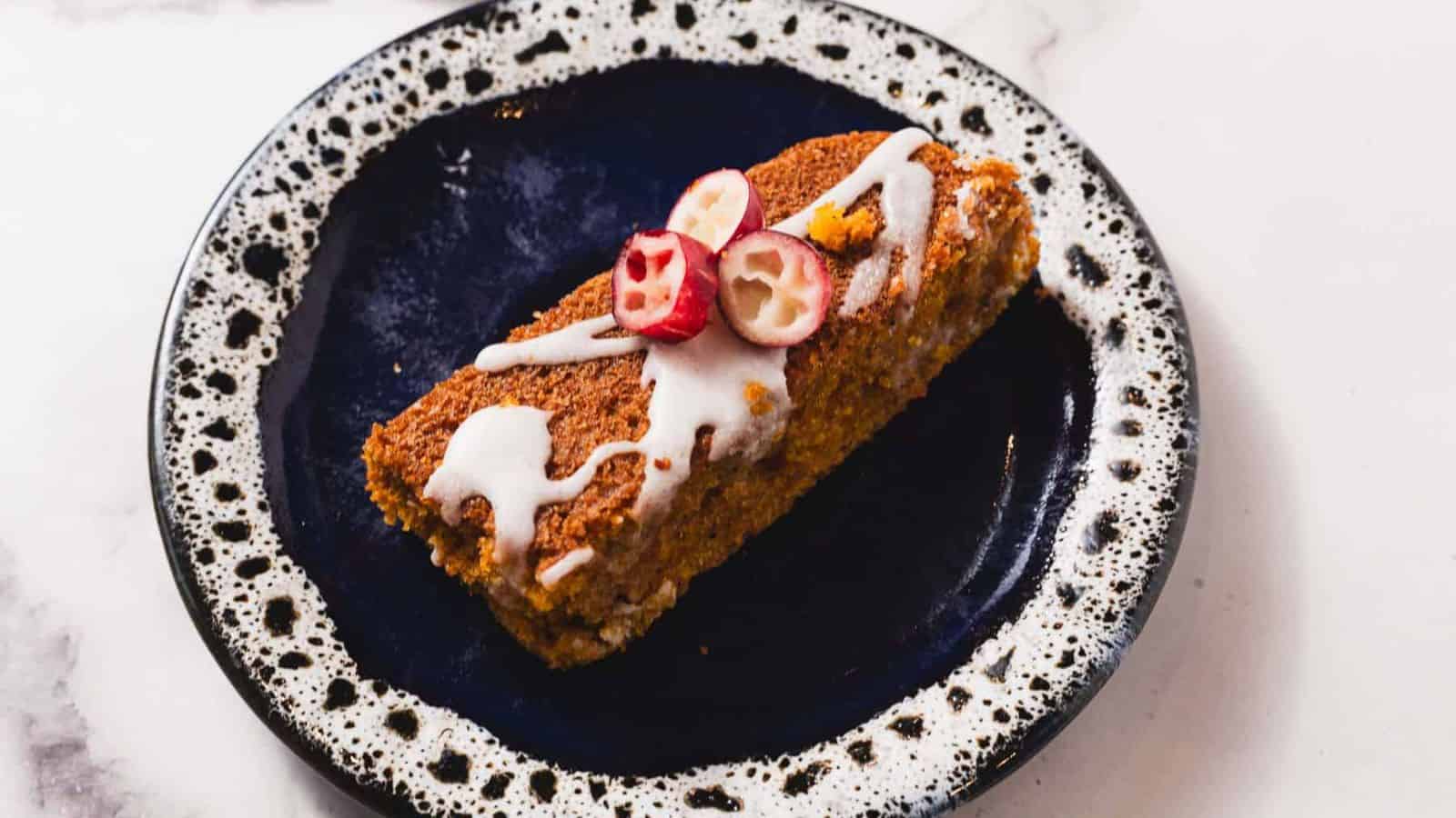 A slice of cake topped with white icing and sliced cranberries is on a black speckled plate with a white rim. The plate is placed on a light-colored marble surface.