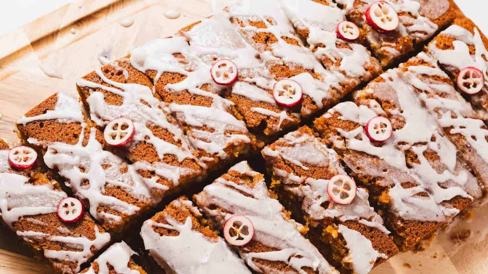 Slices of glazed gingerbread with cranberries on top are arranged on a wooden board. The glaze is drizzled lightly over the surface, contrasting with the rich brown color of the gingerbread. The cranberries are thinly sliced, adding a decorative touch.