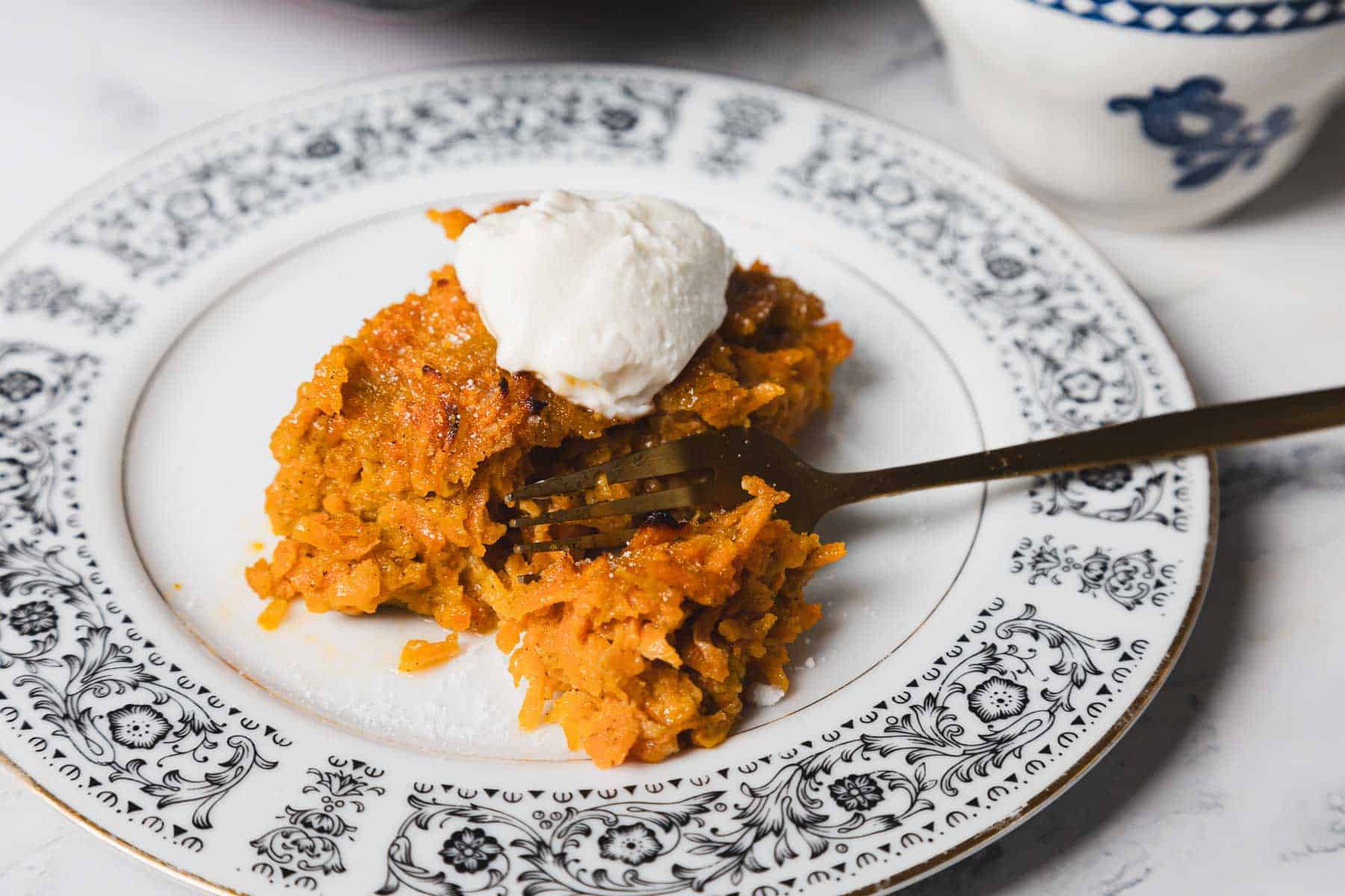 A fork cuts into a slice of carrot pudding topped with a dollop of whipped cream on a white and black floral-patterned plate. A blue and white cup is partially visible in the background.