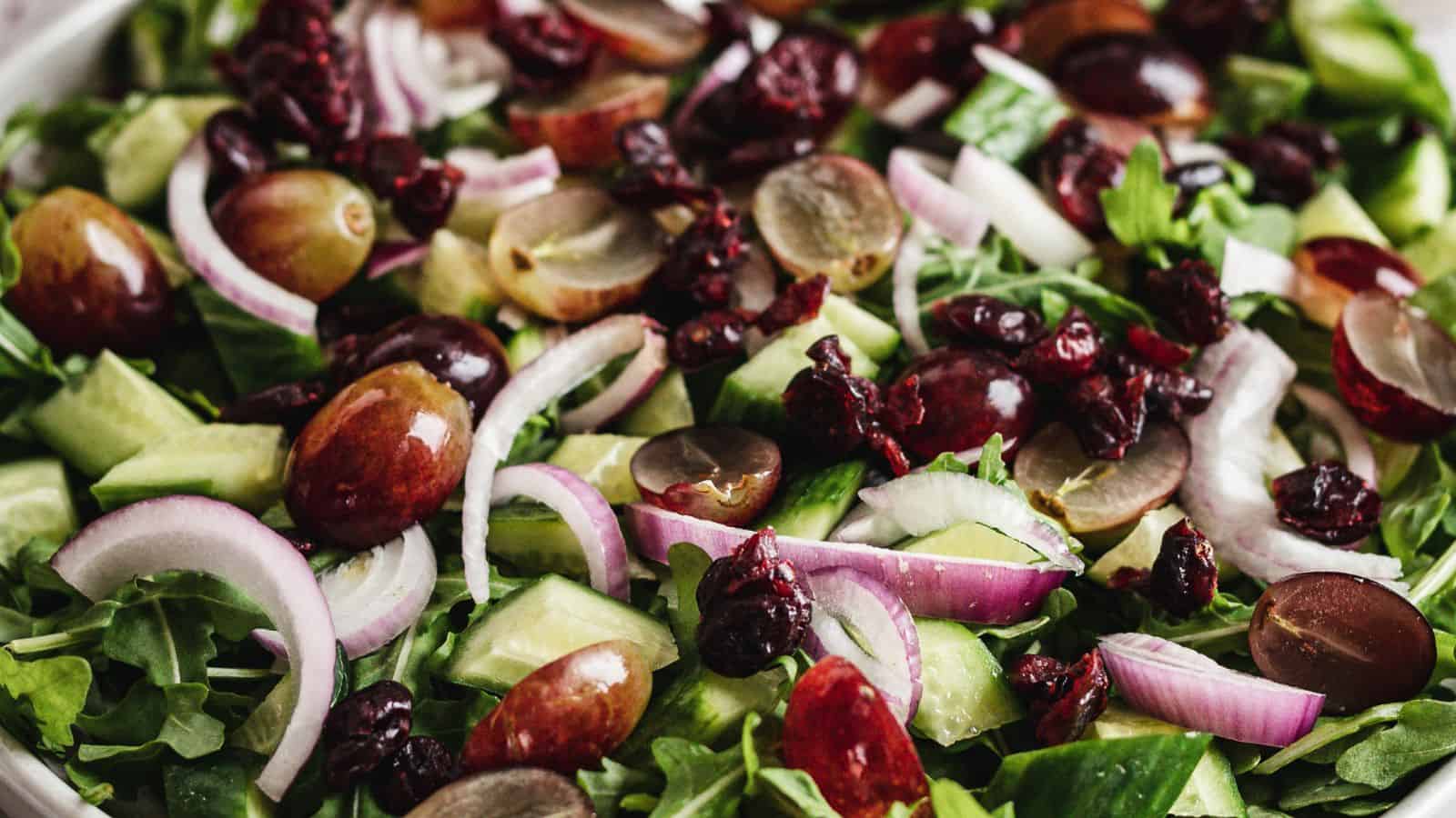 A close-up of a fresh salad featuring sliced grapes, red onions, dried cranberries, cucumber pieces, and a bed of leafy greens. The ingredients are mixed together, showcasing a variety of colors and textures.