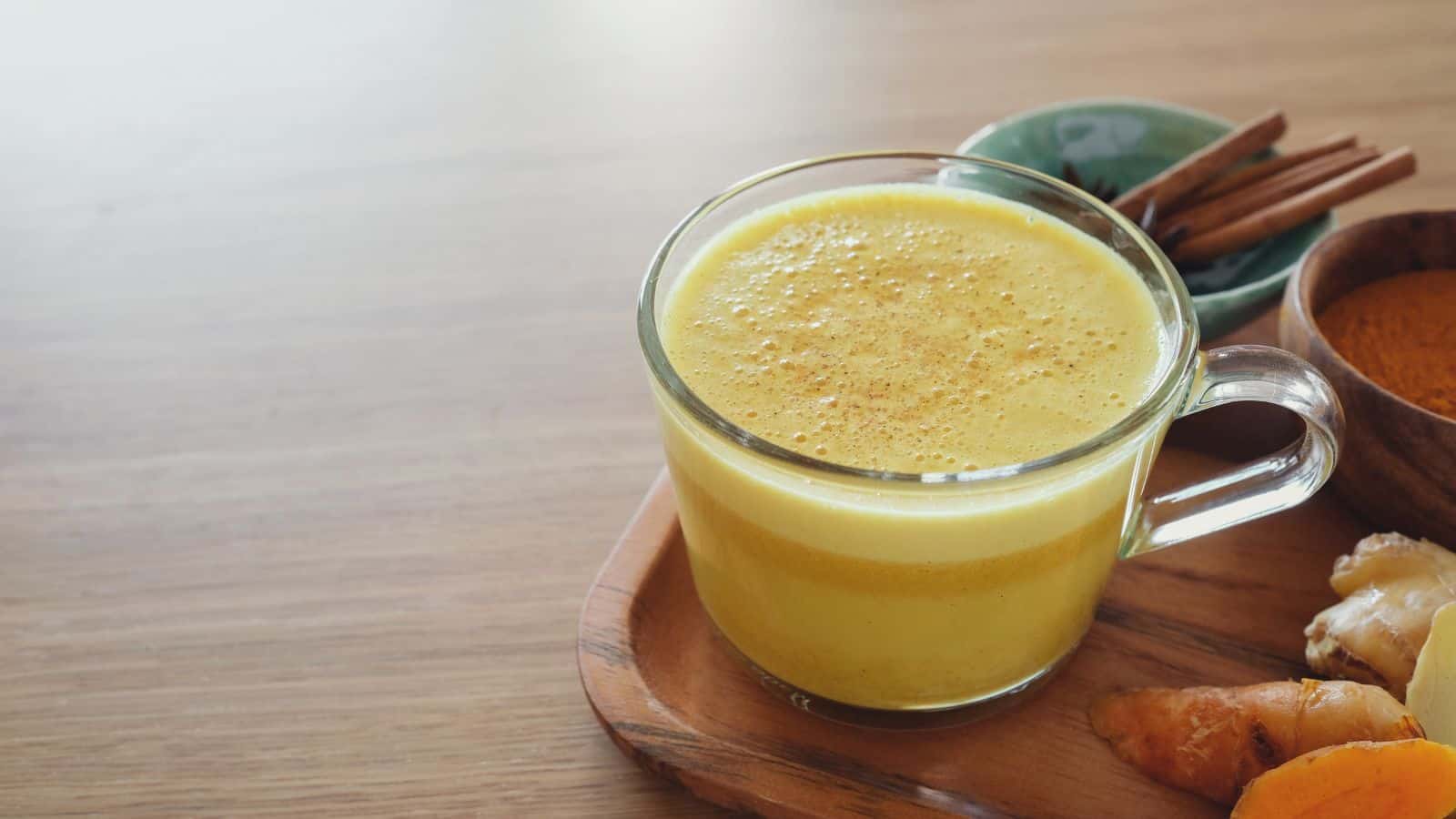 A glass cup filled with yellow turmeric latte is placed on a wooden tray. Behind the cup, there are cinnamon sticks in a small green bowl, a bowl of turmeric powder, and pieces of fresh ginger and turmeric root. The background is a wooden surface.