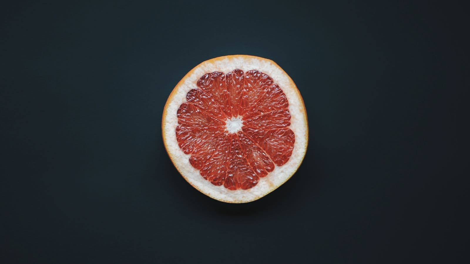 A halved grapefruit with a vibrant red interior and a pale yellow rind is centered against a solid dark background.
