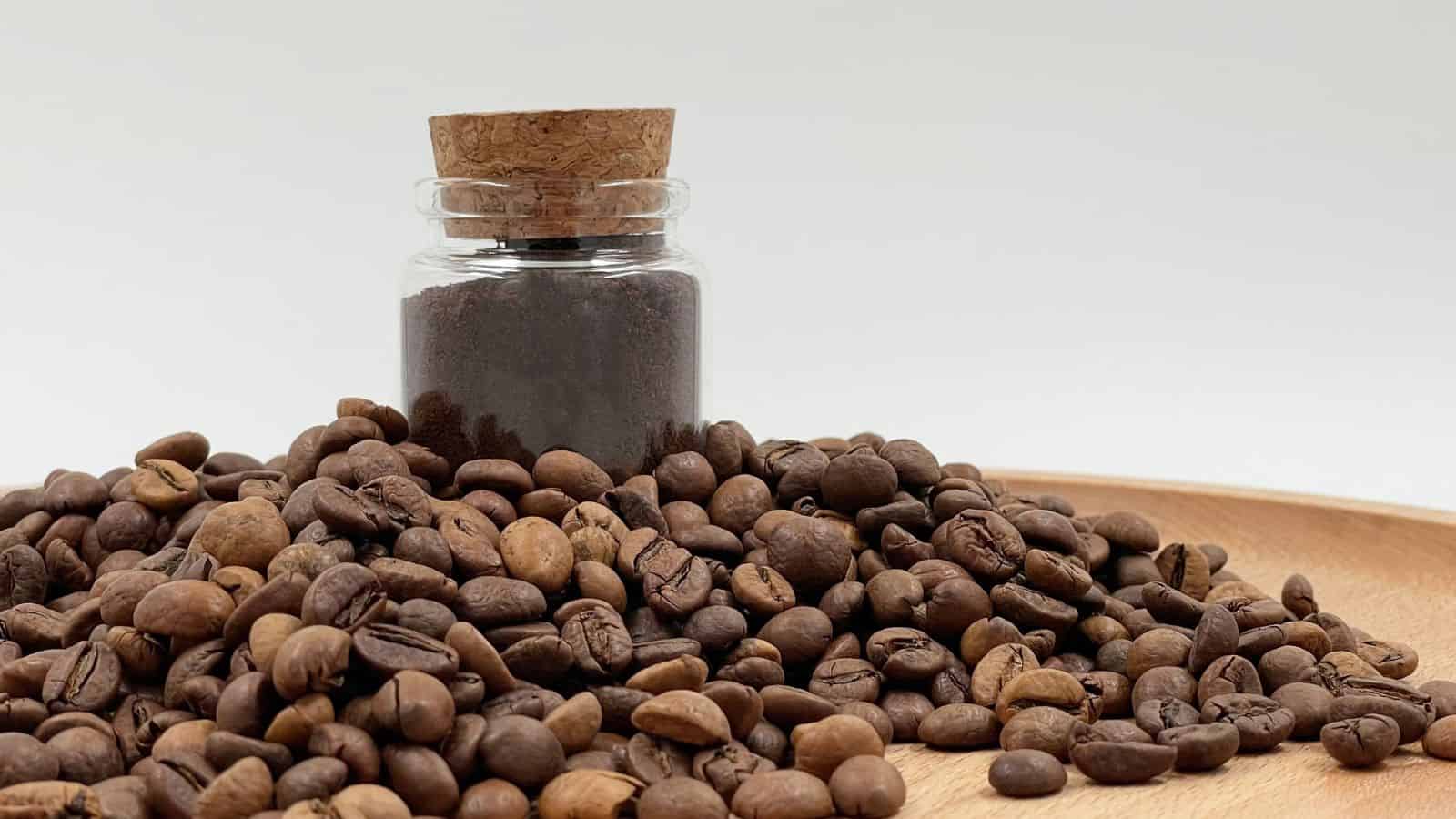 A small glass jar with a cork lid containing ground coffee is placed on a wooden surface. The jar is surrounded by a pile of whole coffee beans.