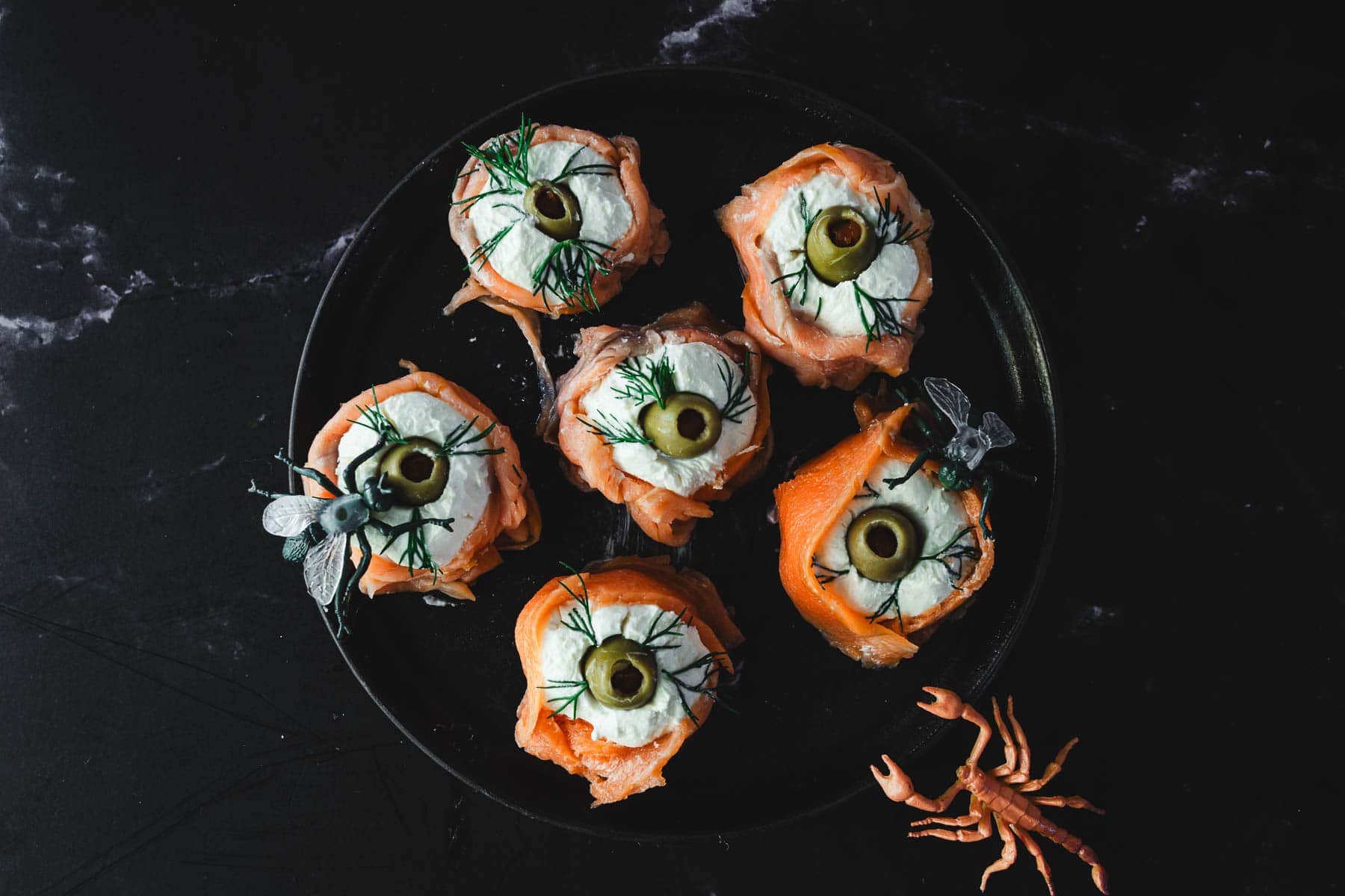 A black plate holds six creatively arranged appetizers resembling eyes, made with cream cheese, olives, and salmon. Dill garnish adds a decorative touch. A small, orange lobster-shaped decoration is placed beside the plate on a dark surface.