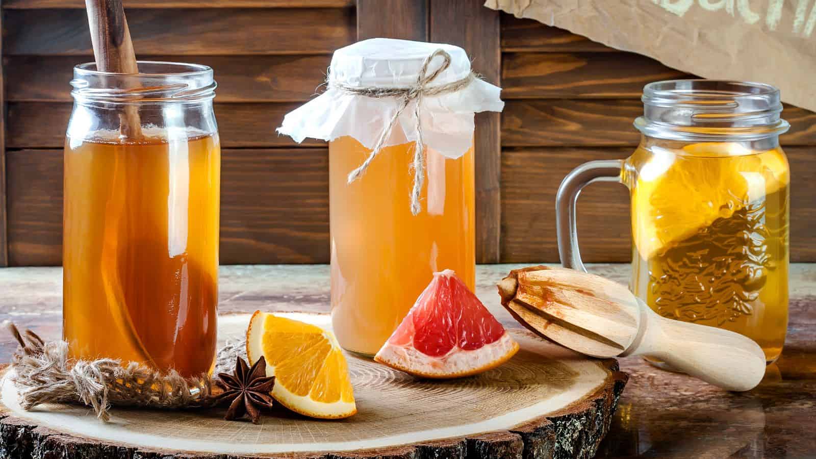 Jars of kombucha are displayed on a wooden surface. One jar has a cinnamon stick, another is covered with cloth and twine, and the third is in a mason jar with a handle. Orange and grapefruit slices, star anise, and a wooden citrus juicer are nearby.
