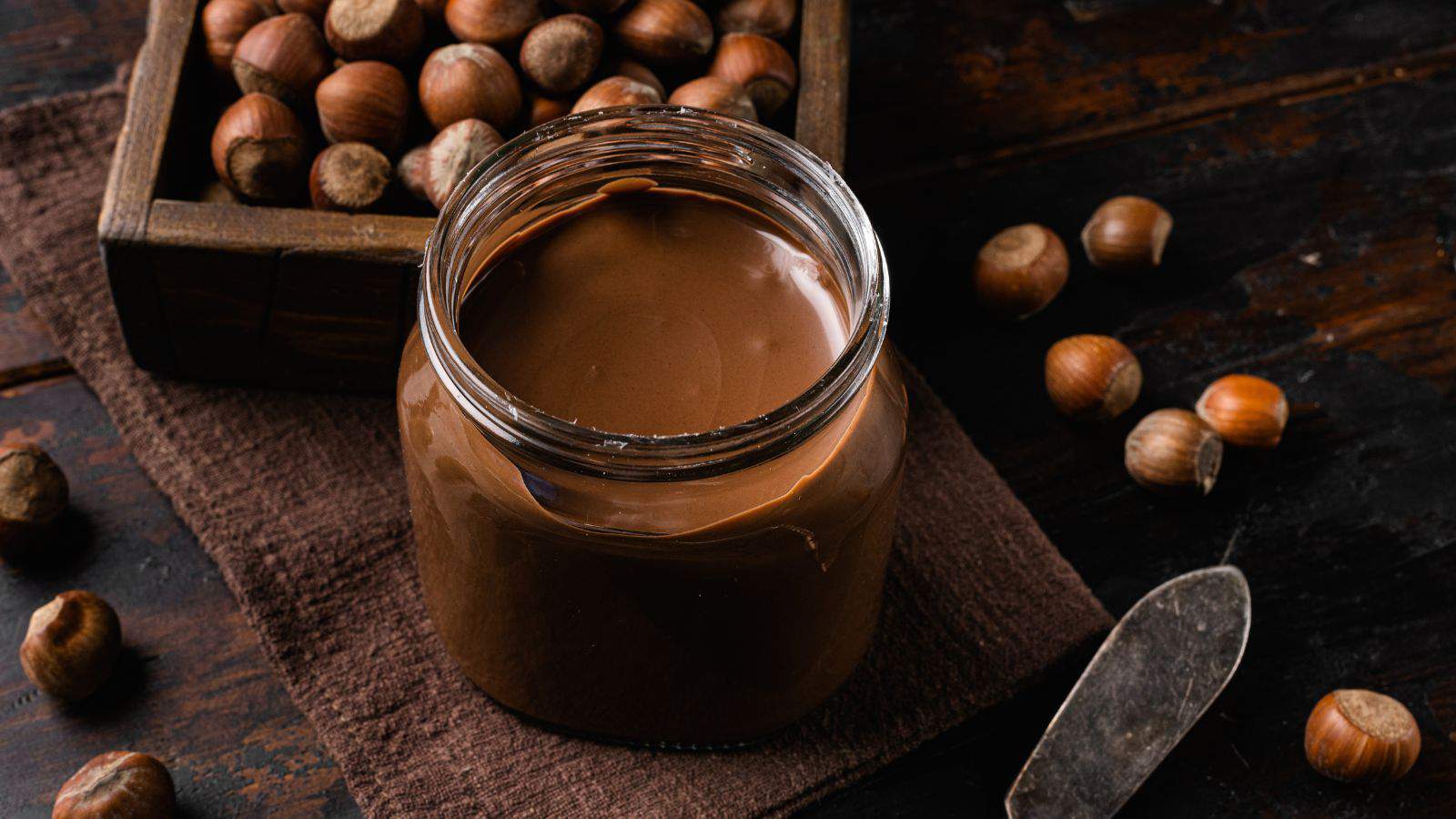 A jar of chocolate hazelnut spread is open on a dark wooden surface. Whole hazelnuts are scattered around, with a wooden box filled with more hazelnuts nearby. A knife rests on a brown cloth beside the jar.