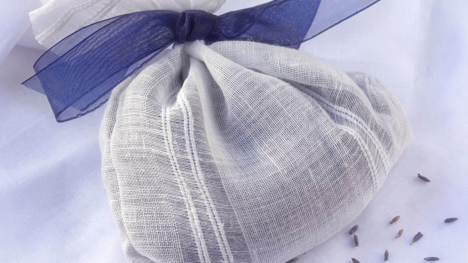 A small lavender sachet made of white fabric with a dark blue ribbon lies on a white surface. A few dried lavender buds are scattered nearby.