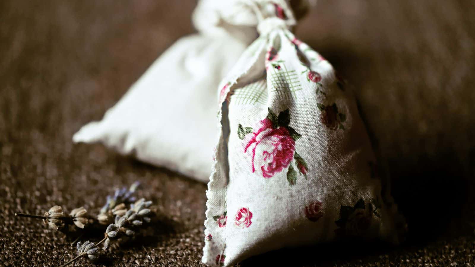 A small fabric pouch with a floral design sits on a textured surface. The pouch is tied at the top. Sprigs of dried lavender rest beside it.