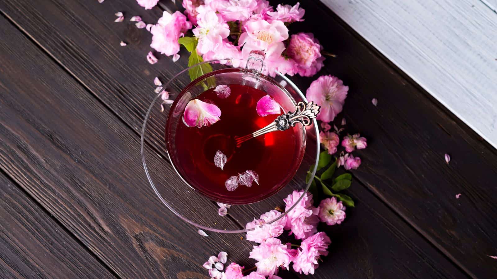A glass cup of red tea with pink flower petals on a saucer and ornate silver spoon, surrounded by scattered pink flowers on a dark wooden table.