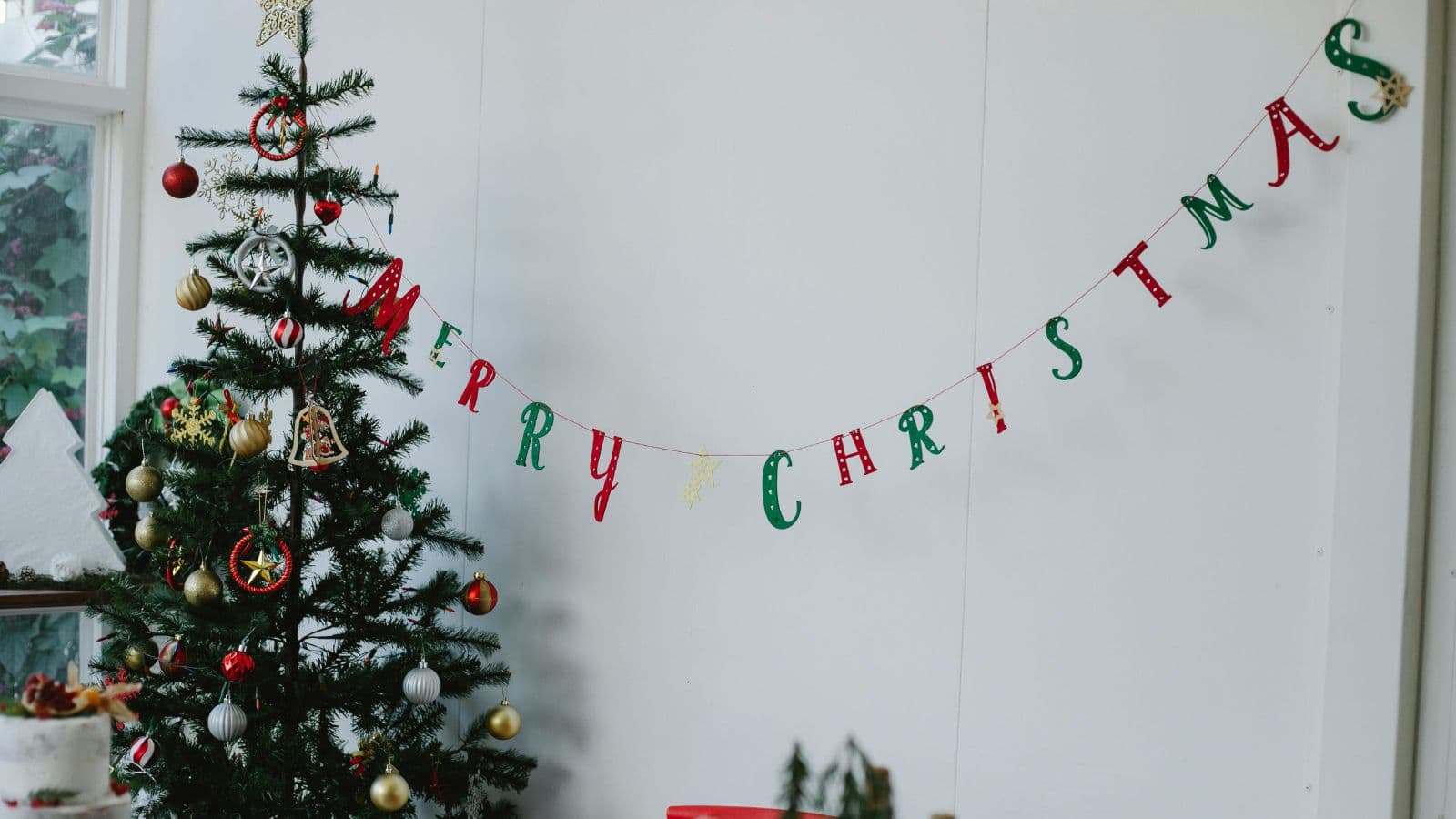 A decorated Christmas tree stands in the corner adorned with ornaments and tinsel. A banner reading "Merry Christmas" is hung on the wall beside the tree. Natural light enters through a nearby window, illuminating the festive scene.