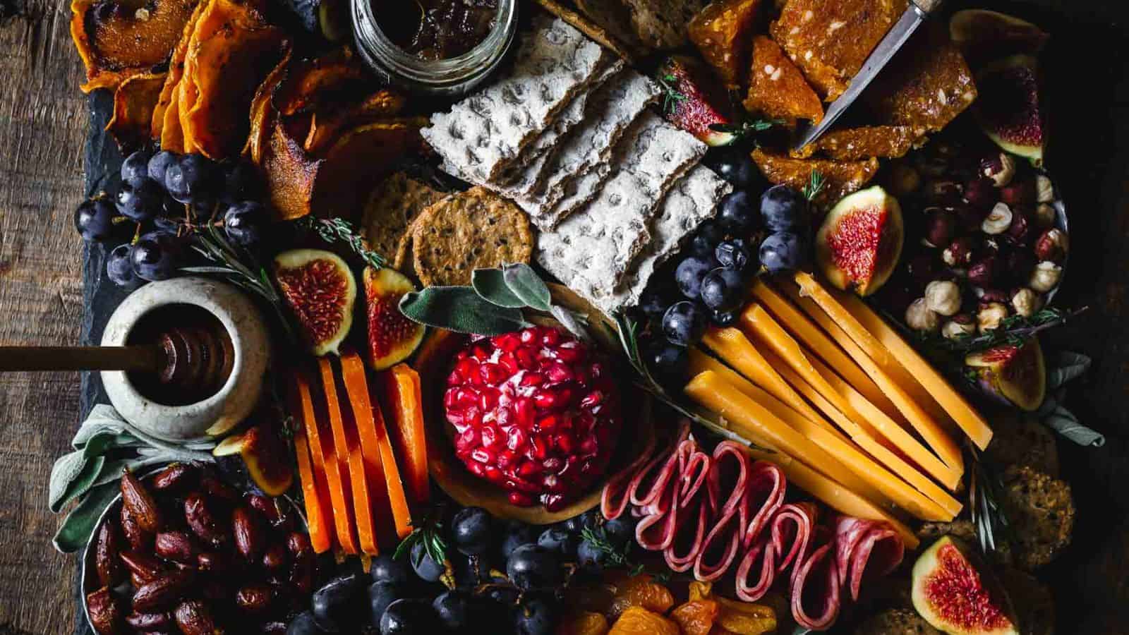 A wooden board with an assortment of cheeses, sliced meats, crackers, and fresh fruits including figs and grapes. There's a central dish with pomegranate seeds, a jar of preserves, and a bowl with honey alongside a honey dipper.