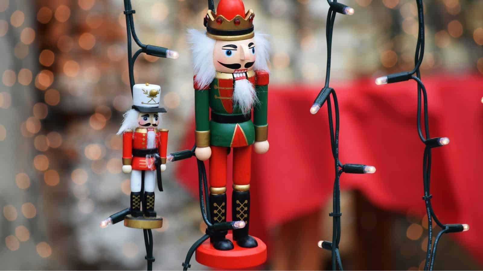 Two nutcracker figurines, one large and one small, are displayed among hanging string lights. The background features blurred bokeh lighting and a red surface.