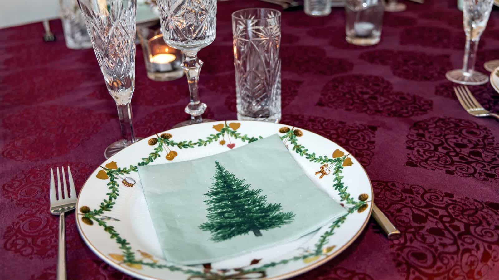A dining table set with a white plate featuring a Christmas tree napkin. Surrounding cutlery includes a fork and knife. Crystal glassware and a lit candle are on a red patterned tablecloth.