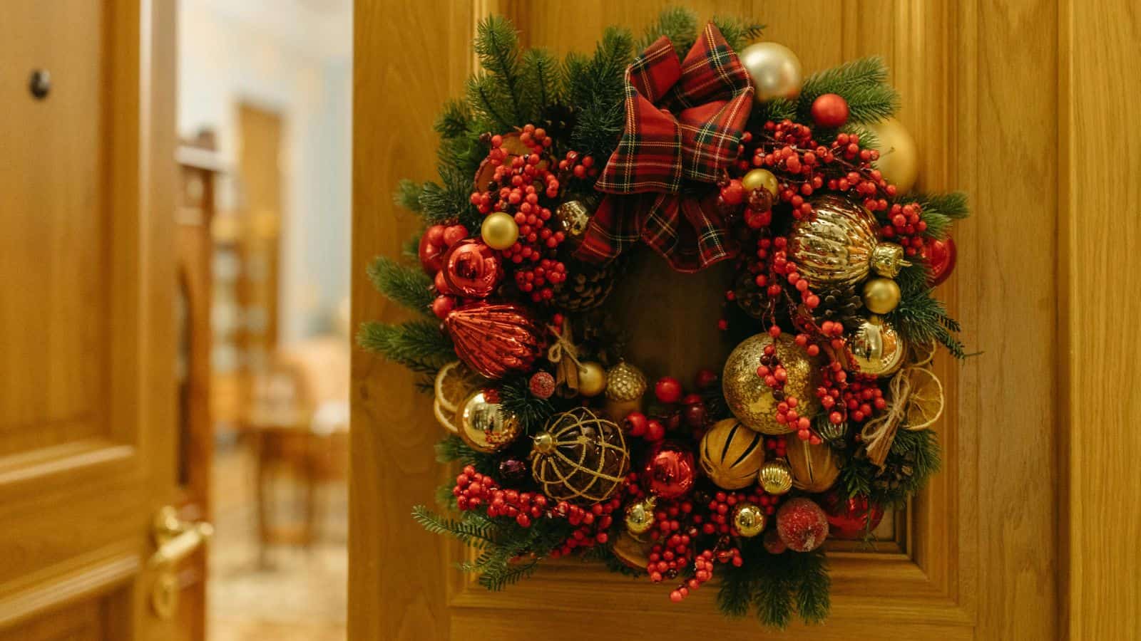 A festive Christmas wreath hangs on a wooden door. The wreath is adorned with red and gold ornaments, pine cones, and a red tartan bow at the top.