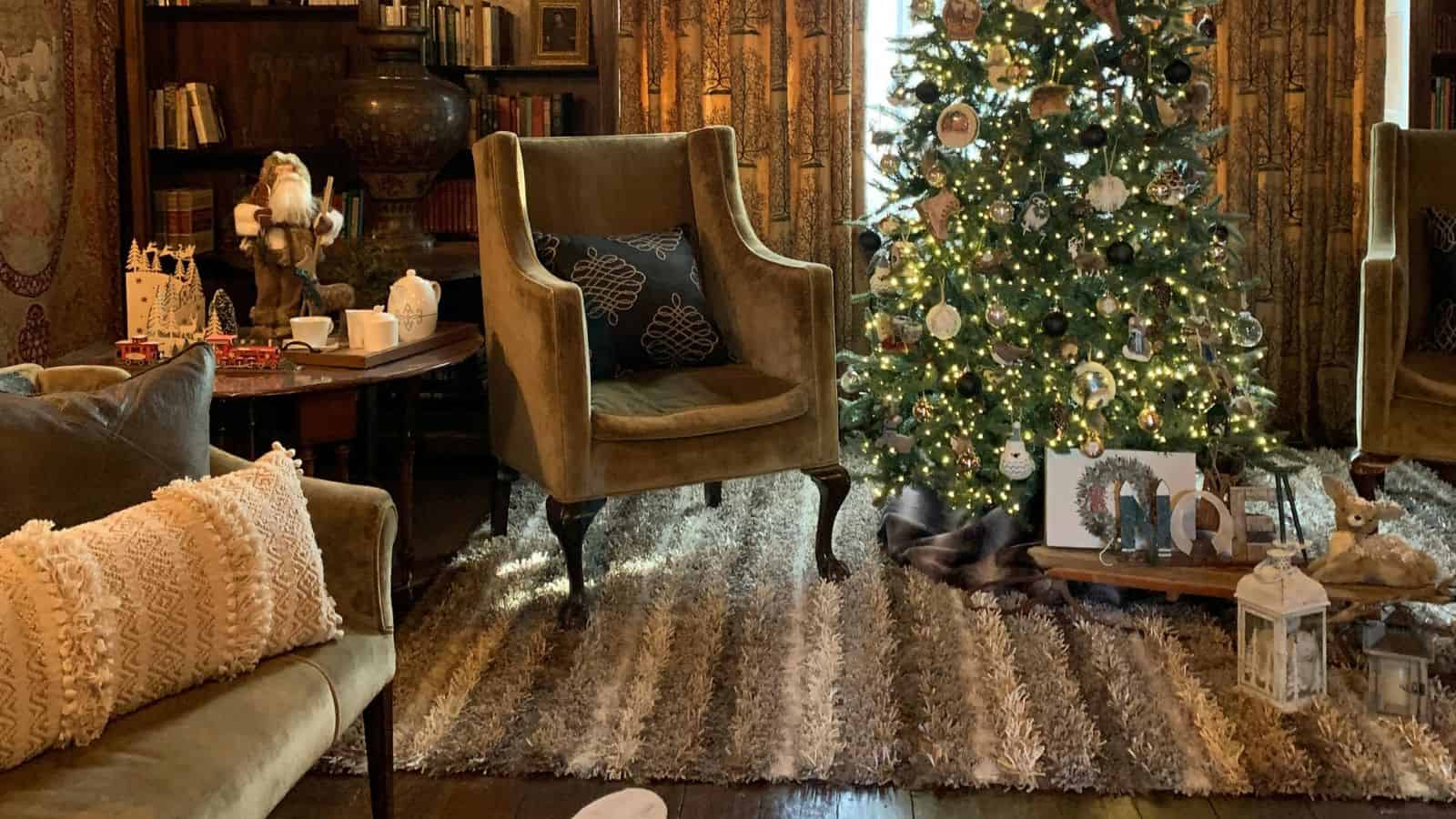 A cozy living room with a decorated Christmas tree on a patterned rug. An armchair and a sofa with cushions are nearby. A small table holds festive decorations and a Santa figurine. Shelves with books are in the background.