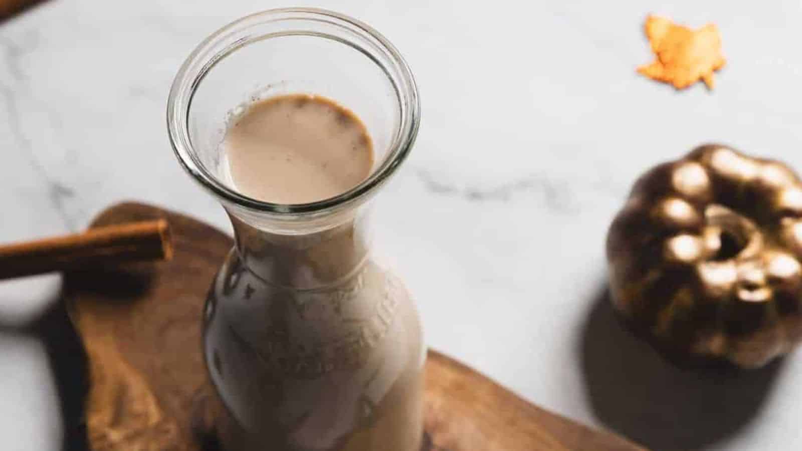 A glass bottle filled with a creamy liquid sits on a wooden board. A cinnamon stick is visible on the left. In the background, there is a small pumpkin decoration and an orange leaf on a marble surface.