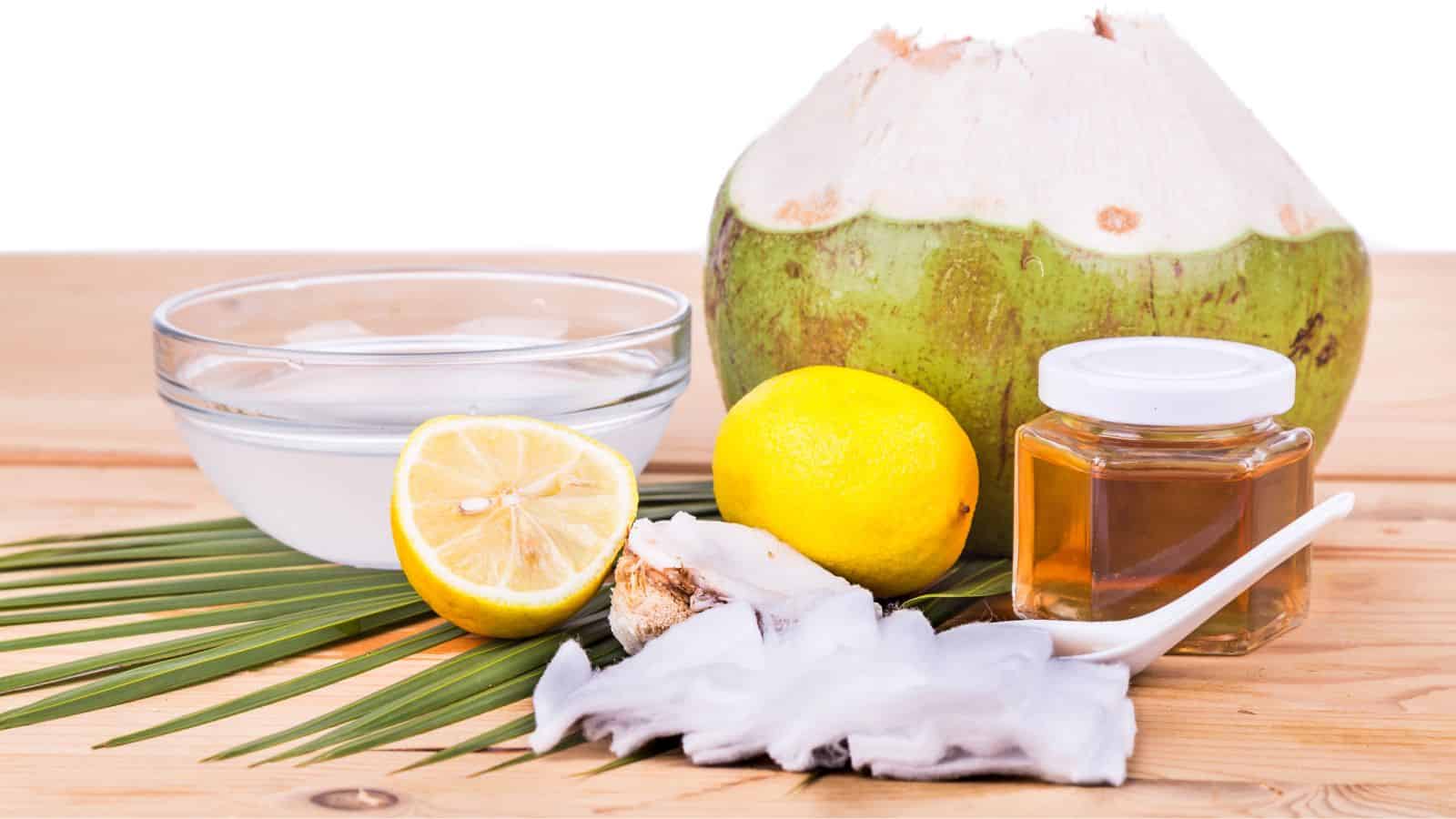 Coconut, bowl of coconut water, cut lemon, whole lemon, a jar of honey, and a spoon with coconut meat on a wooden surface. Palm leaf placed underneath.