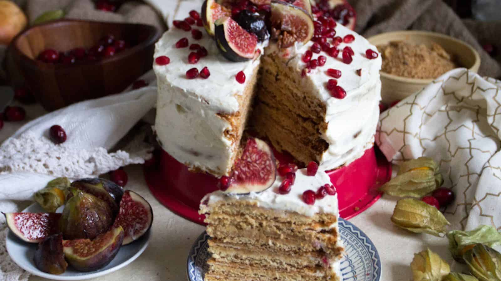 A multi-layered cake with white frosting is topped with figs, pomegranate seeds, and nuts. A slice is cut and placed on a plate. Surrounding the cake are physalis, sliced figs, and a bowl of brown sugar. A white cloth is draped nearby.