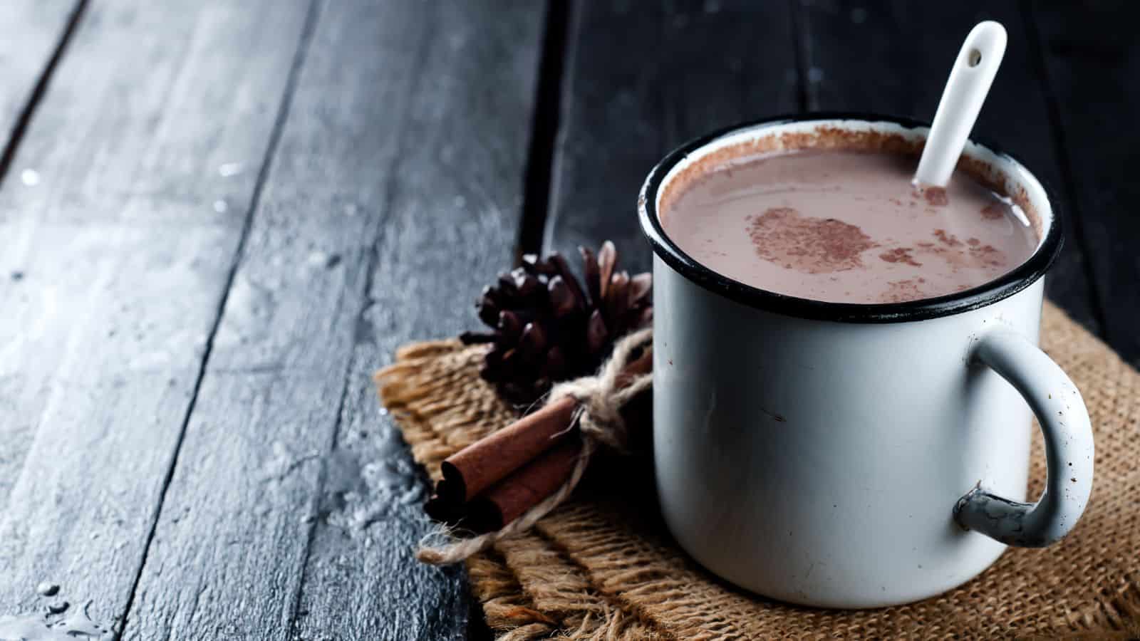 A white ceramic mug filled with hot chocolate sits on a piece of burlap on a dark wooden table. A spoon is placed inside the mug. Cinnamon sticks and a pine cone are situated next to the mug, adding a rustic touch.