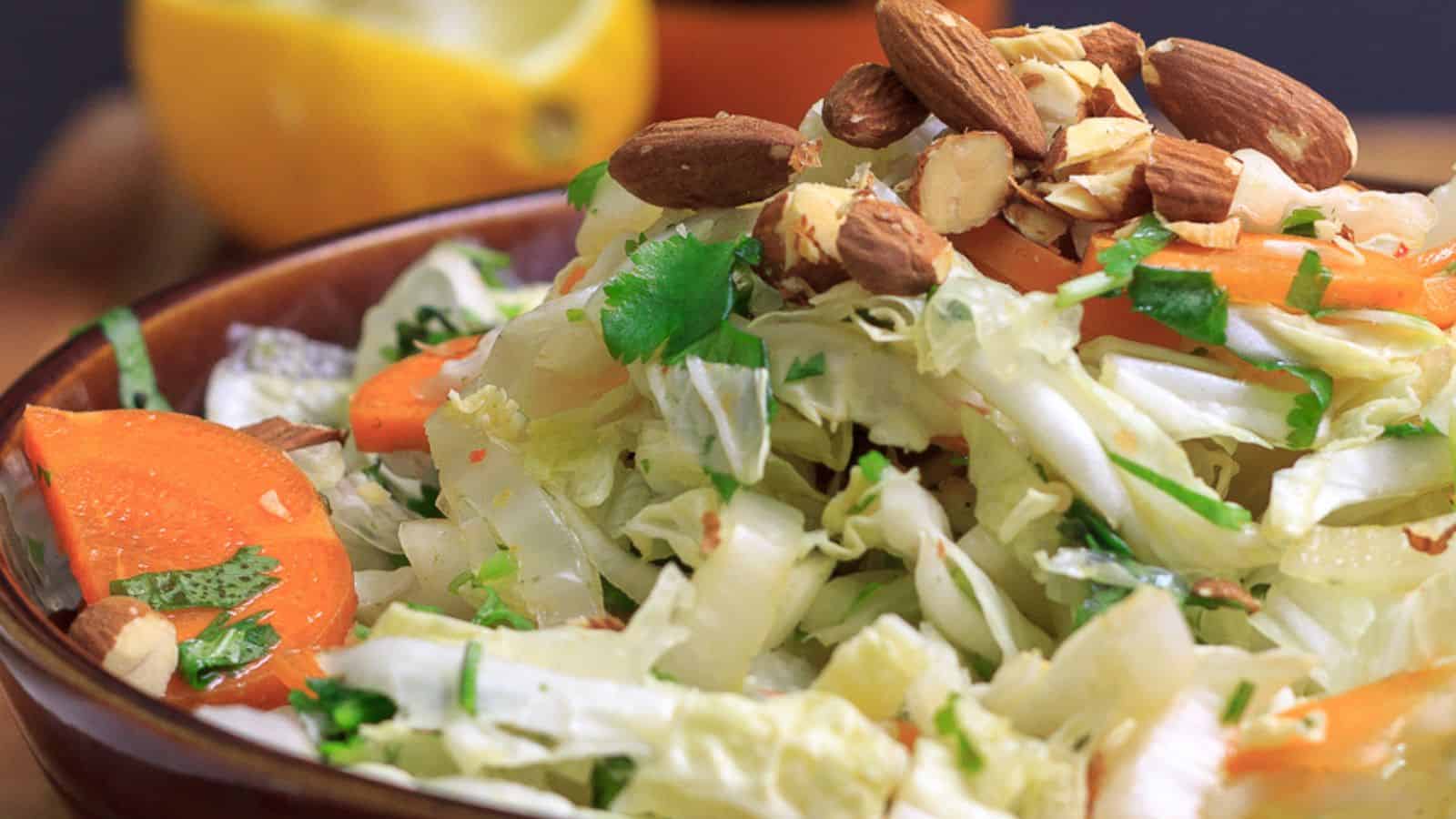 A close-up of a salad featuring chopped cabbage, sliced carrots, cilantro, and whole almonds. In the background, there is a partially squeezed lemon on a wooden surface.