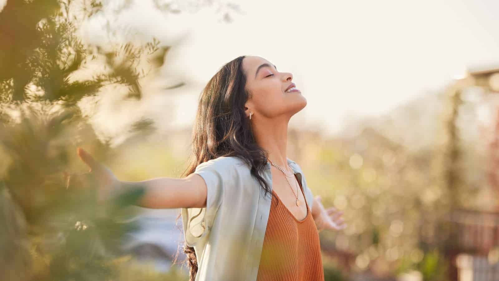 A person with long hair stands outdoors with eyes closed, arms outstretched, enjoying the sunlight. The background is blurred with greenery and soft natural light, creating a serene atmosphere.