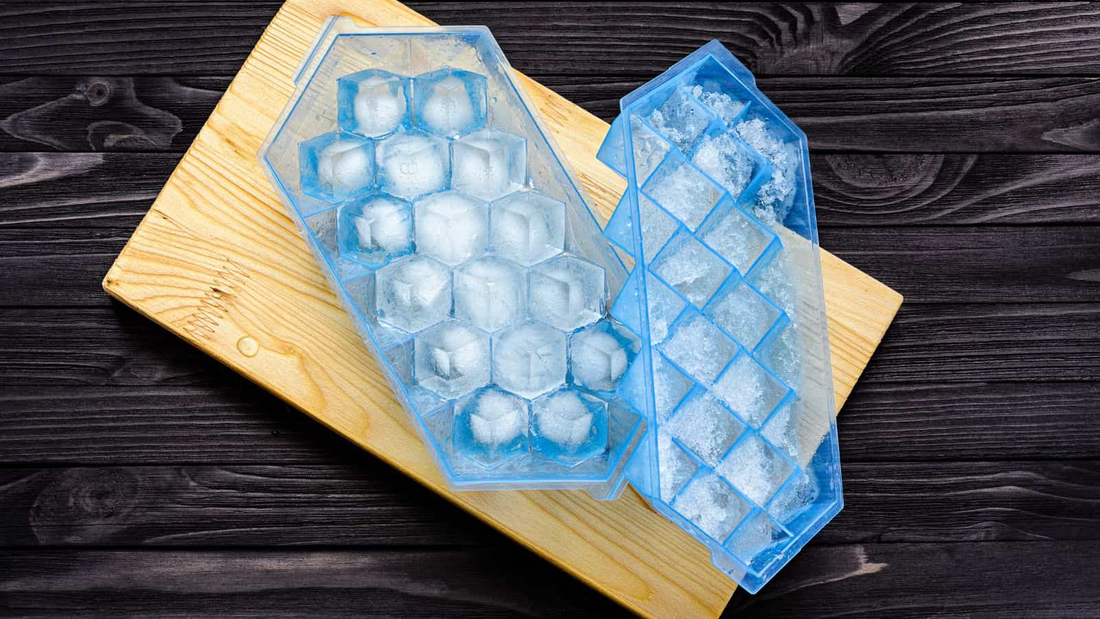 Two blue silicone ice cube trays, one filled with clear hexagonal ice cubes, rest on a wooden cutting board with a dark wood background. The second tray is empty, showing frost and ice remnants.