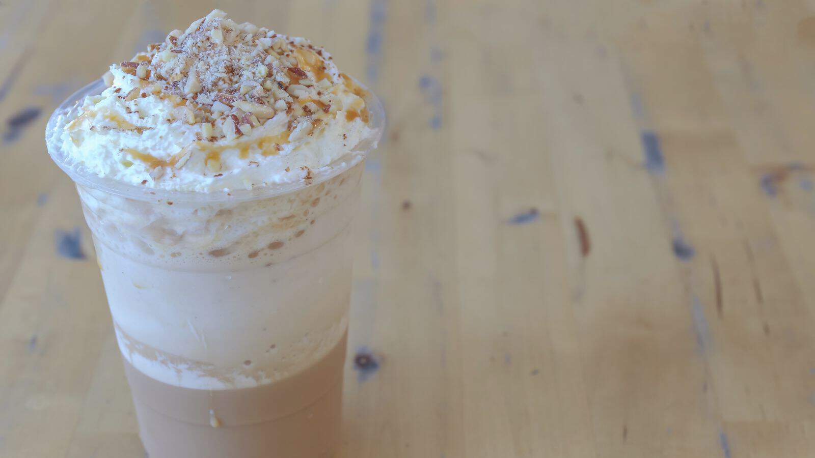 A plastic cup filled with a creamy beverage topped with whipped cream, caramel drizzle, and sprinkled nuts. The drink is placed on a light wooden surface.