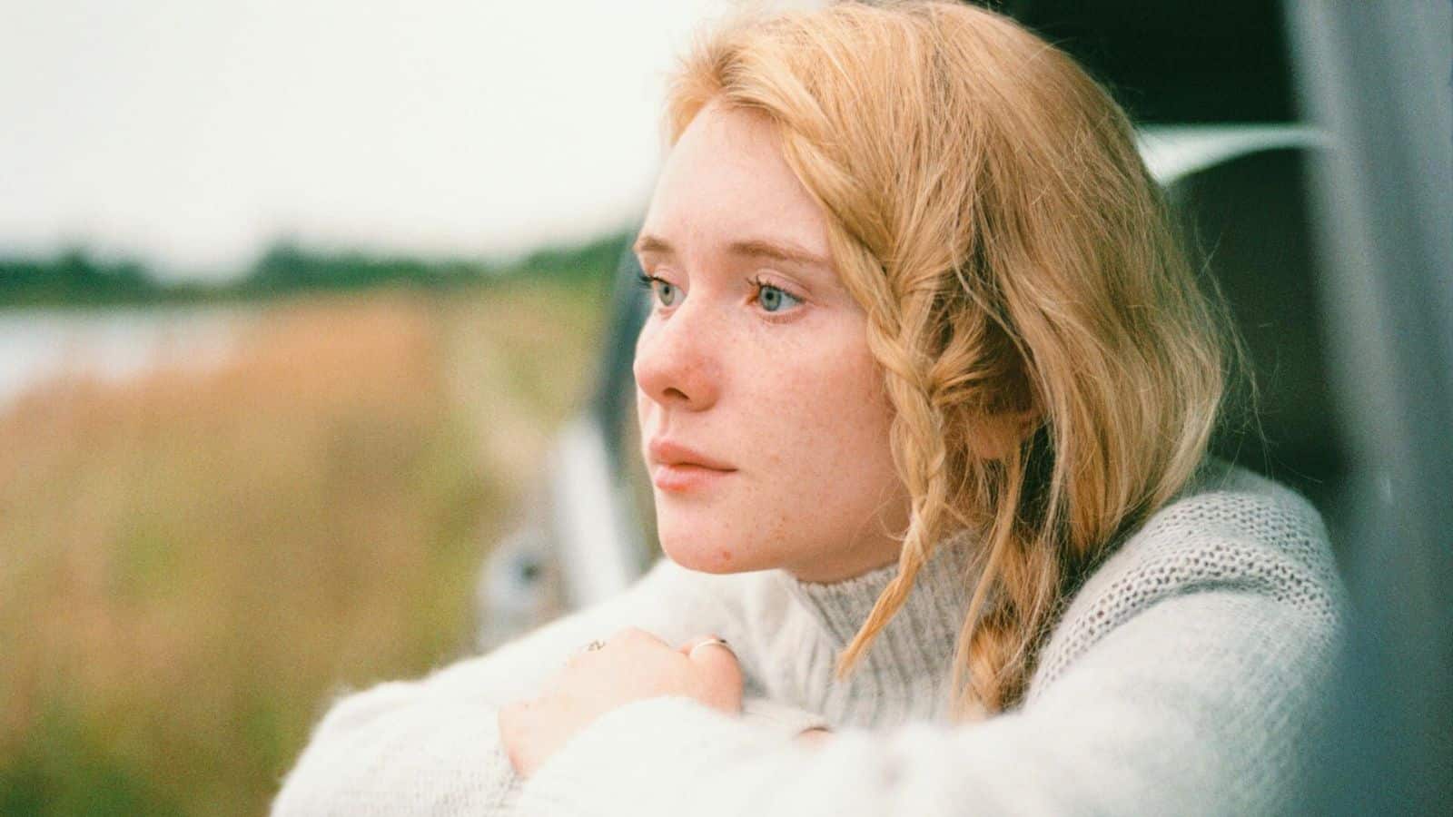 A person with long, light-colored hair and a pensive expression gazes out of a car window. They wear a light sweater. The background shows a blurred view of grass and sky, suggesting a rural or countryside setting.