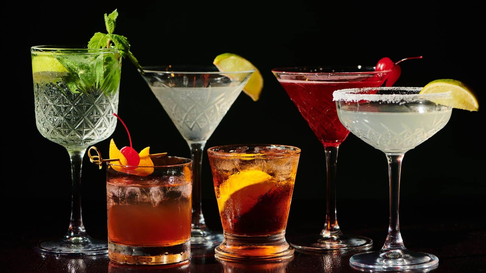 A variety of six cocktails displayed against a dark background. Each glass contains a different beverage garnished with elements like lime, cherries, mint, and a sugared rim, showcasing assorted colors and textures.