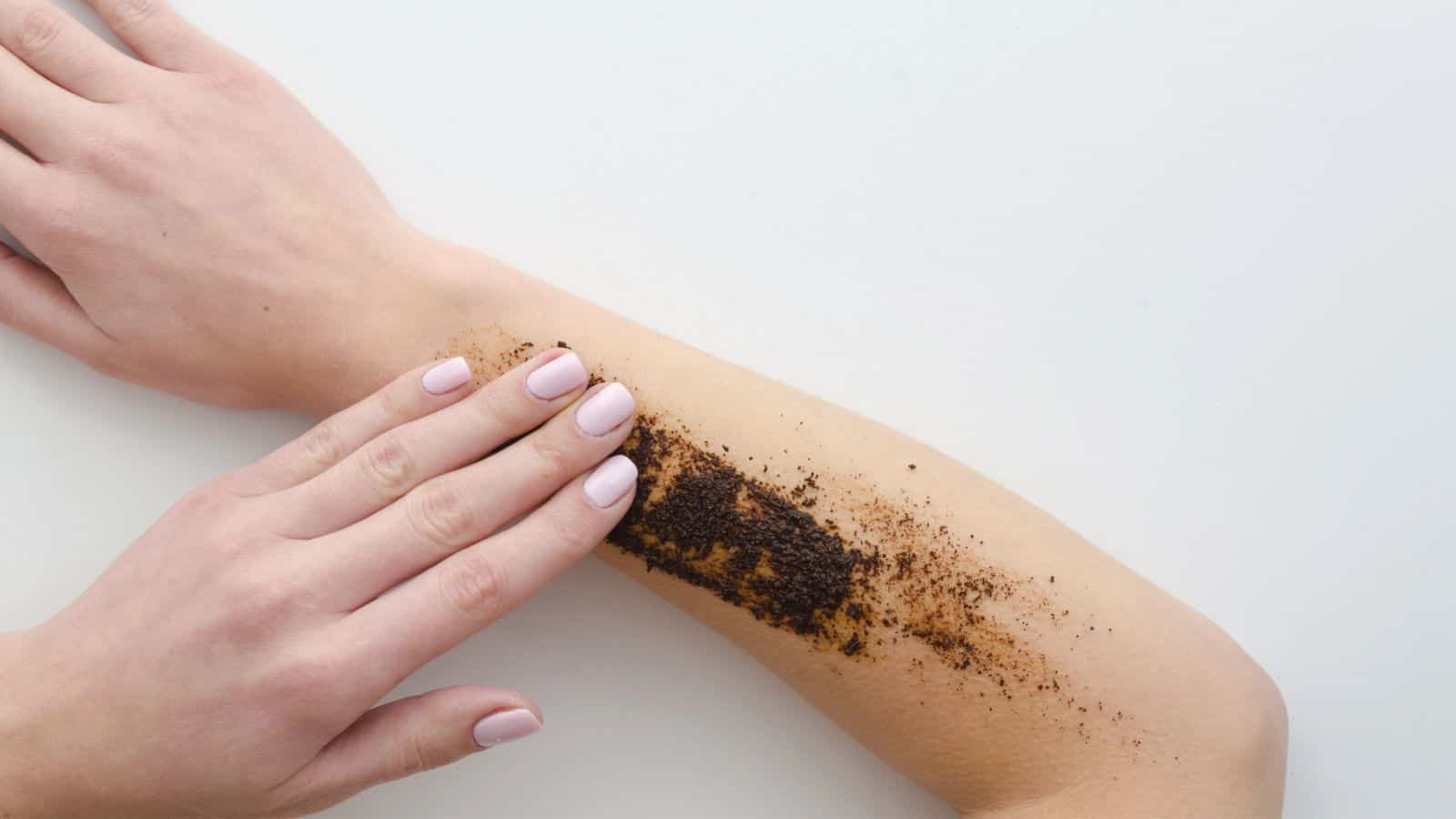 A person explores surprising coffee grounds uses by applying them to their forearm with their fingers. The grounds form a small rectangular patch on the skin, against the backdrop of a plain white surface.