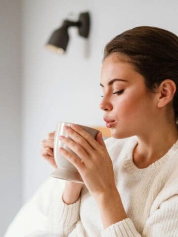 A woman with brown hair in a ponytail is sitting on a bed, holding and smelling a white mug. She is wearing a white sweater, and a black wall-mounted light fixture is visible in the background.