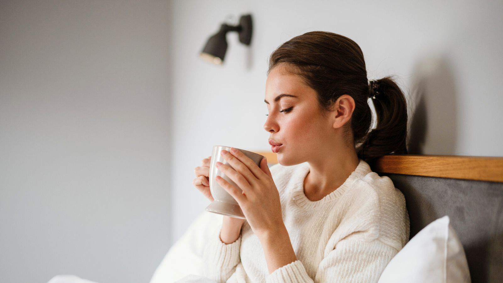 A woman with brown hair in a ponytail is sitting on a bed, holding and smelling a white mug. She is wearing a white sweater, and a black wall-mounted light fixture is visible in the background.