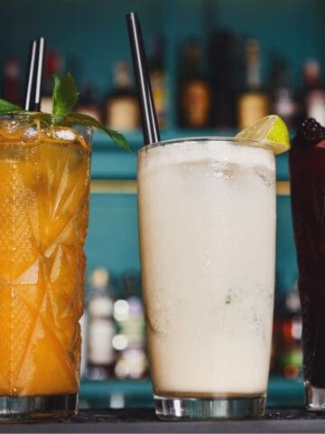 Three cocktails on a bar counter: one orange with mint leaves, one creamy white with a lime wedge, and one dark red with a leafy garnish, each in a textured glass with a black straw. Background shows an array of blurred bottles.