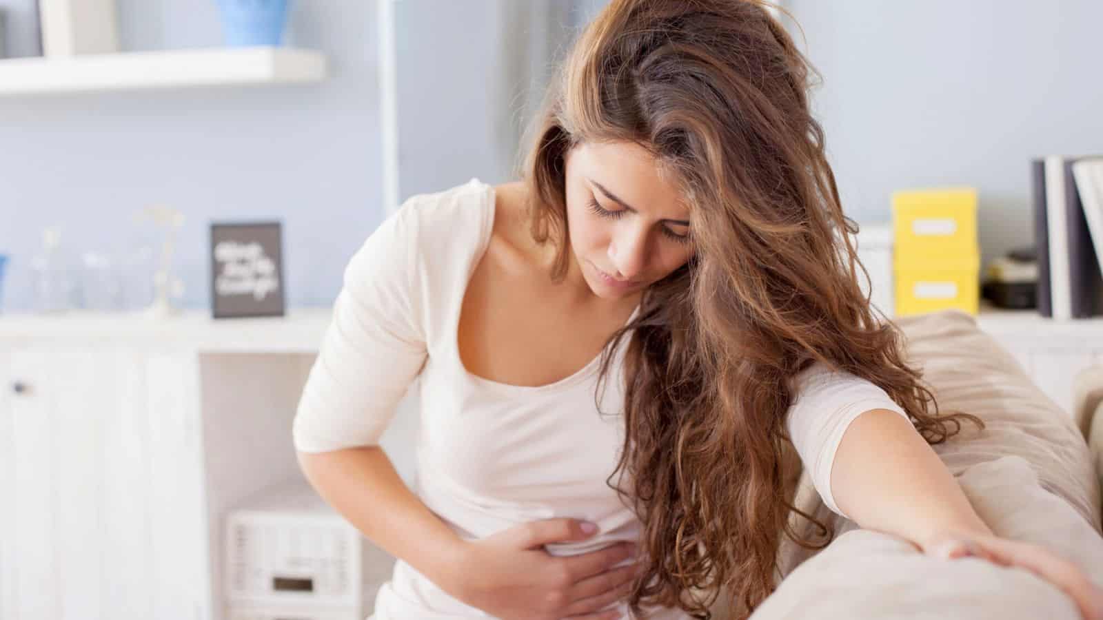 A woman with long brown hair, wearing a white top, is sitting on a couch. She is holding her stomach with one hand and leaning on the couch with the other. The background includes a shelf with a few decorative items.