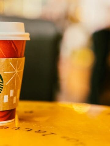 A Starbucks paper cup with a lid and cardboard sleeve sits on a yellow table. The cup features a red design and the Starbucks logo. The background is blurred, showing indistinct shapes and colors.