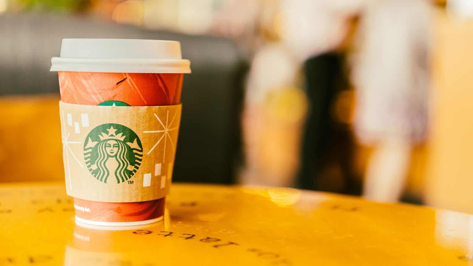 A Starbucks paper cup with a lid and cardboard sleeve sits on a yellow table. The cup features a red design and the Starbucks logo. The background is blurred, showing indistinct shapes and colors.