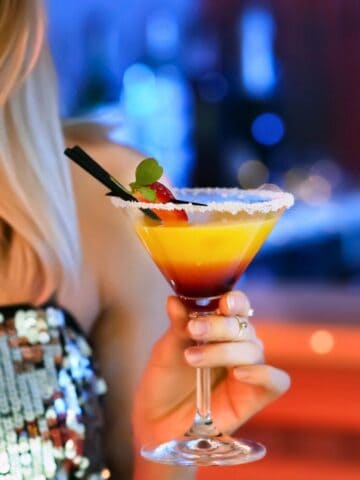 A person wearing a sequined top holds a cocktail in a stem glass. The drink has layers of yellow and red, garnished with a straw and small fruit. The background is softly blurred, with hints of a bar setting.