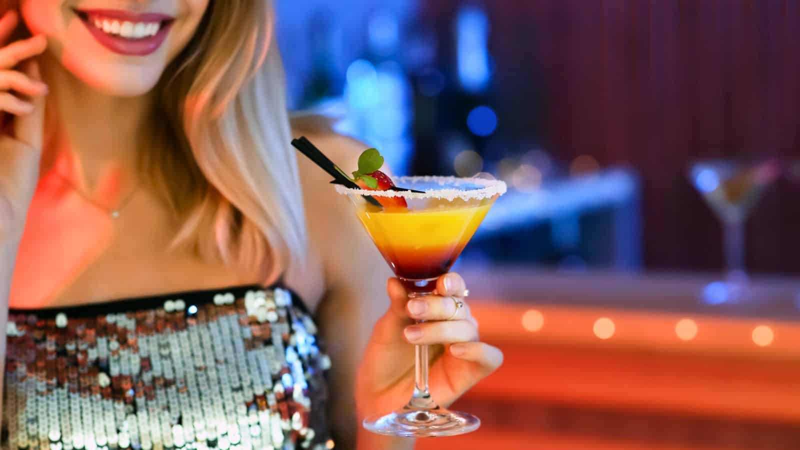A person wearing a sequined top holds a cocktail in a stem glass. The drink has layers of yellow and red, garnished with a straw and small fruit. The background is softly blurred, with hints of a bar setting.