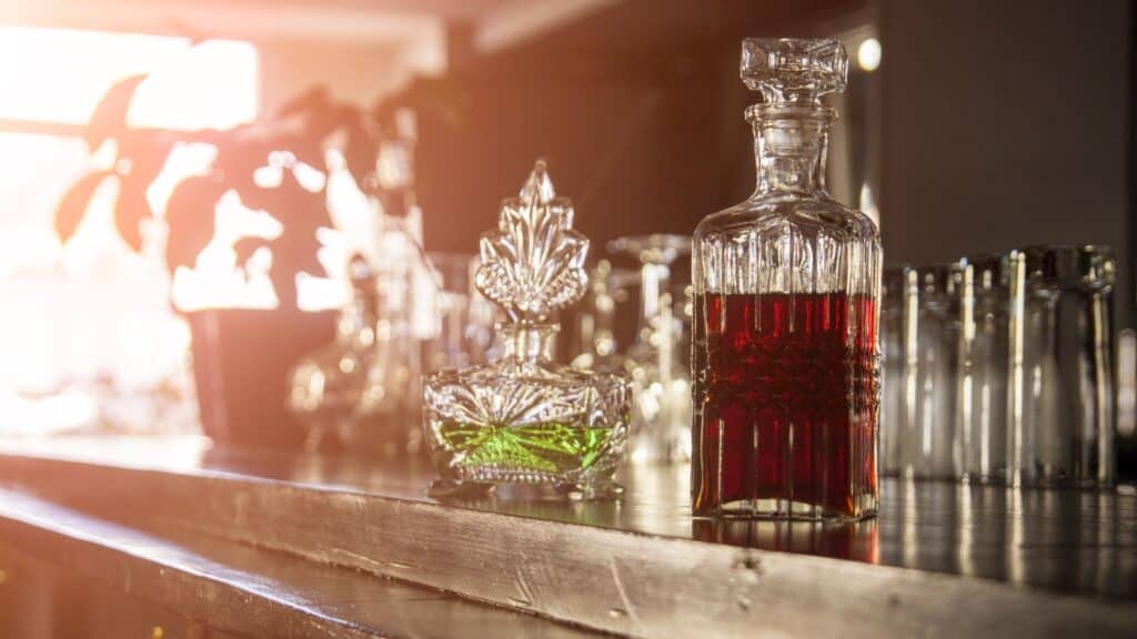 Two ornate decanters sit on a wooden bar counter, perfect for your holiday drink station. The left decanter holds a green liquid with a pointed stopper, while the right one contains red liquid and features a square-shaped stopper. Sunlight filters in, creating a warm ambiance.