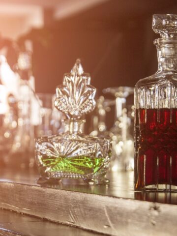 Two ornate decanters sit on a wooden bar counter, perfect for your holiday drink station. The left decanter holds a green liquid with a pointed stopper, while the right one contains red liquid and features a square-shaped stopper. Sunlight filters in, creating a warm ambiance.