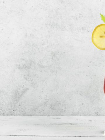 A tall glass of Bloody Mary, garnished with celery, a lemon slice, a cherry tomato, and a pickle spear, standing against a light gray background.